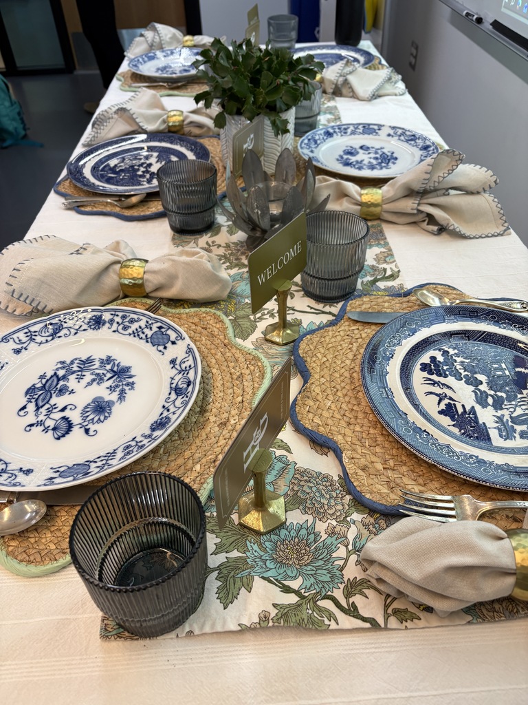 Elegant table setting with a white tablecloth, blue-and-white china plates with floral and pastoral designs, and gold-accented beige napkins held in gold rings. Woven placemats and ribbed dark gray glasses add texture. A small sign reads “WELCOME,” and a centerpiece features green foliage in a gray vase. The table runner has a floral pattern in green, blue, and brown tones.