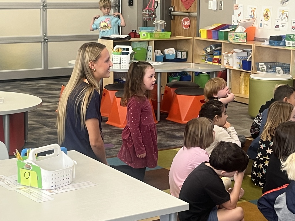 A young female student teacher kneels next to a smiling kindergartener in a maroon dress as they interact with a seated group of children in a colorful classroom. A young boy stands in the background near a play kitchen.