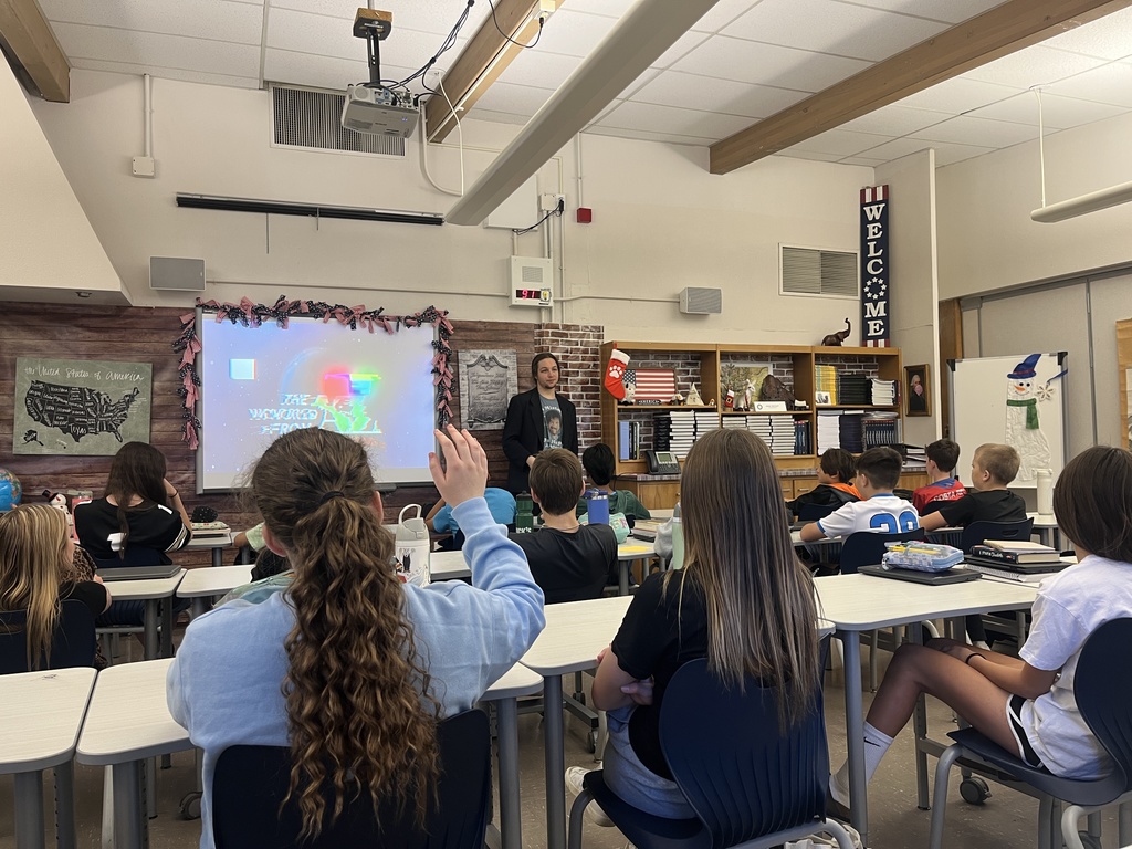 A classroom of upper elementary students listens to a guest speaker at the front of the room, who stands beside a screen displaying the title “The Wayward Real From Paul.” A student in the front row raises their hand.