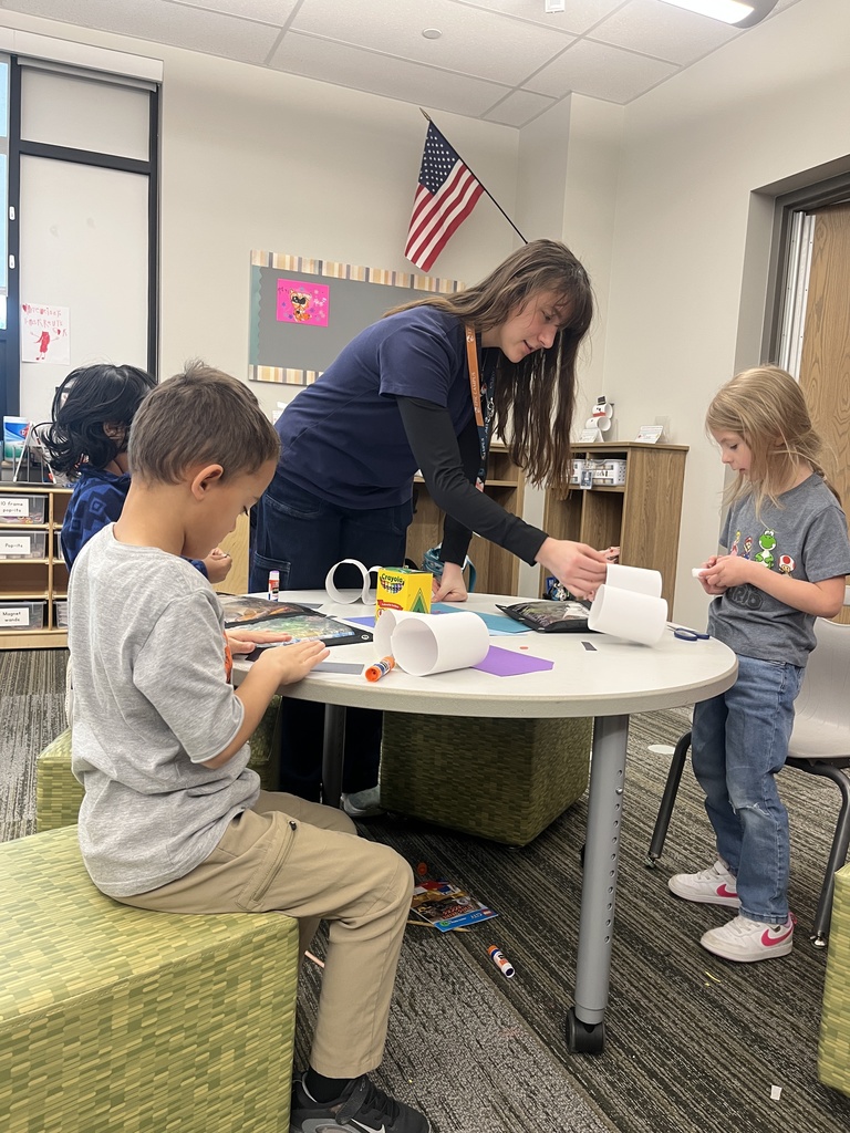 A teacher works with three young students at a small table in a classroom. The children are cutting and gluing paper for a craft activity. An American flag hangs in the background.