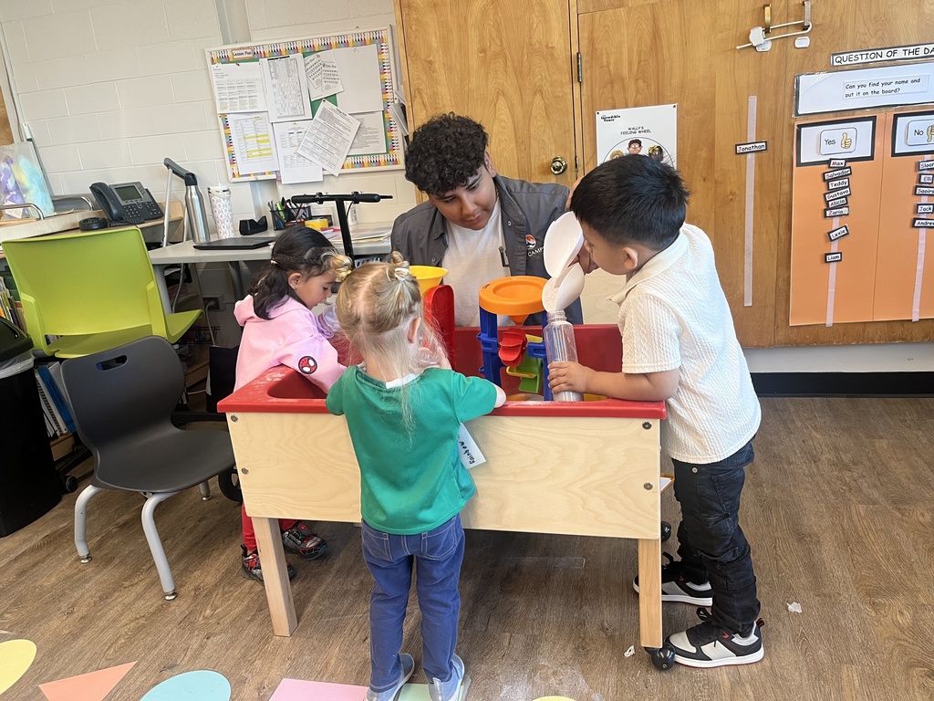 A group of four young children, two girls and two boys of diverse backgrounds, play at a sensory table filled with funnels and tubes in a colorful classroom. An older student or adult supervises, seated behind them. The room includes charts, bulletin boards, and labeled classroom materials.