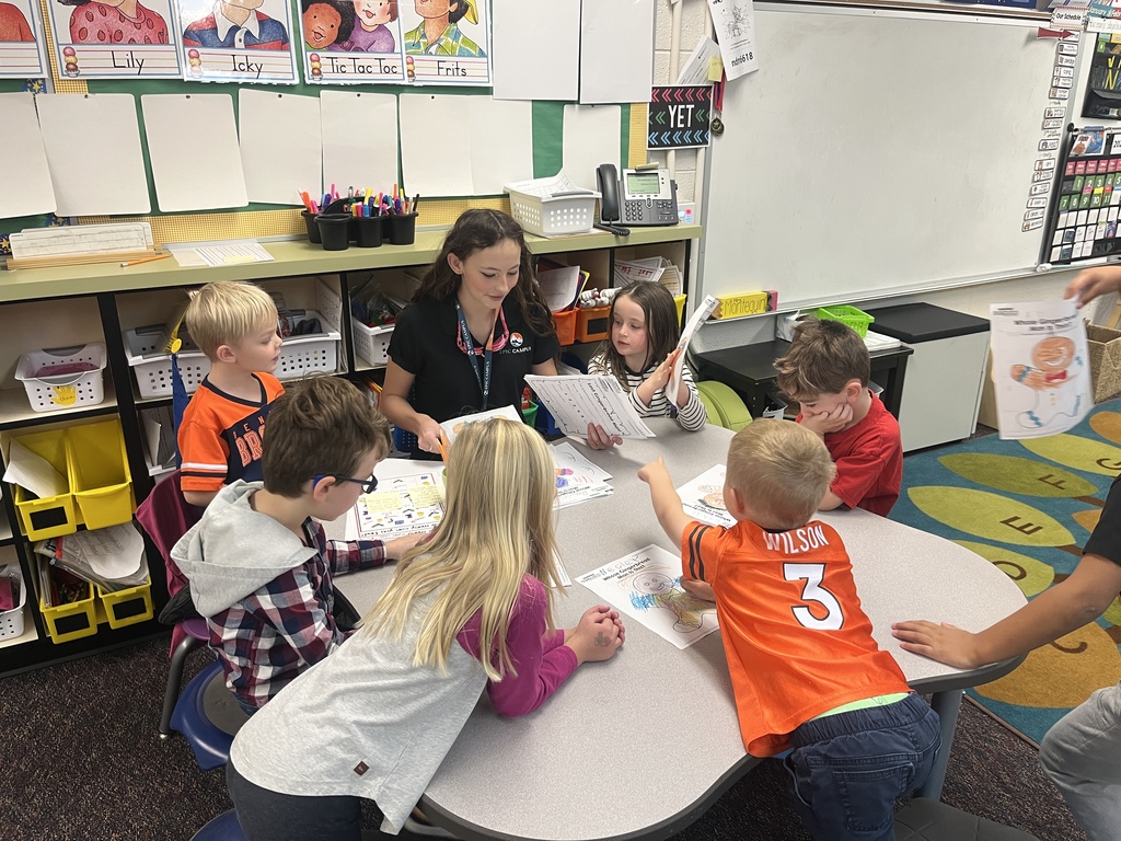 A group of young students gathers around a table, working on activity sheets with a young female teacher wearing an EPIC Campus lanyard. The classroom is colorful, with student artwork and name cards displayed.