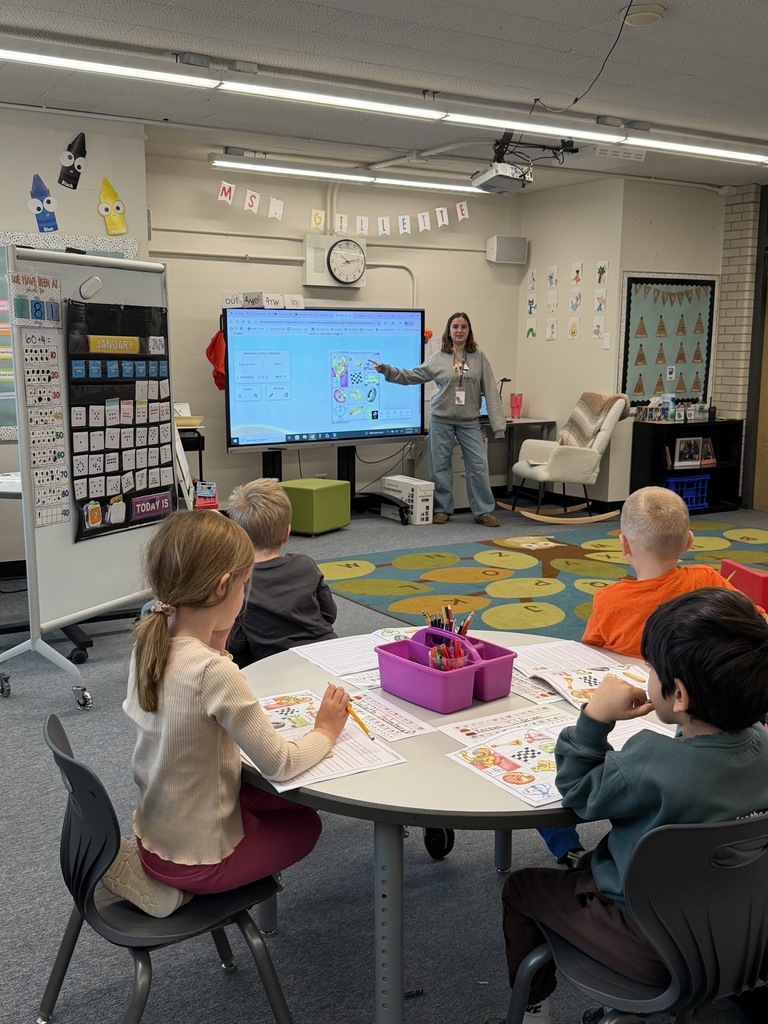 A teacher stands at the front of a classroom pointing to a large digital screen while young students at round tables follow along with worksheets. The room includes colorful decor, banners, and educational displays like a calendar and number chart.