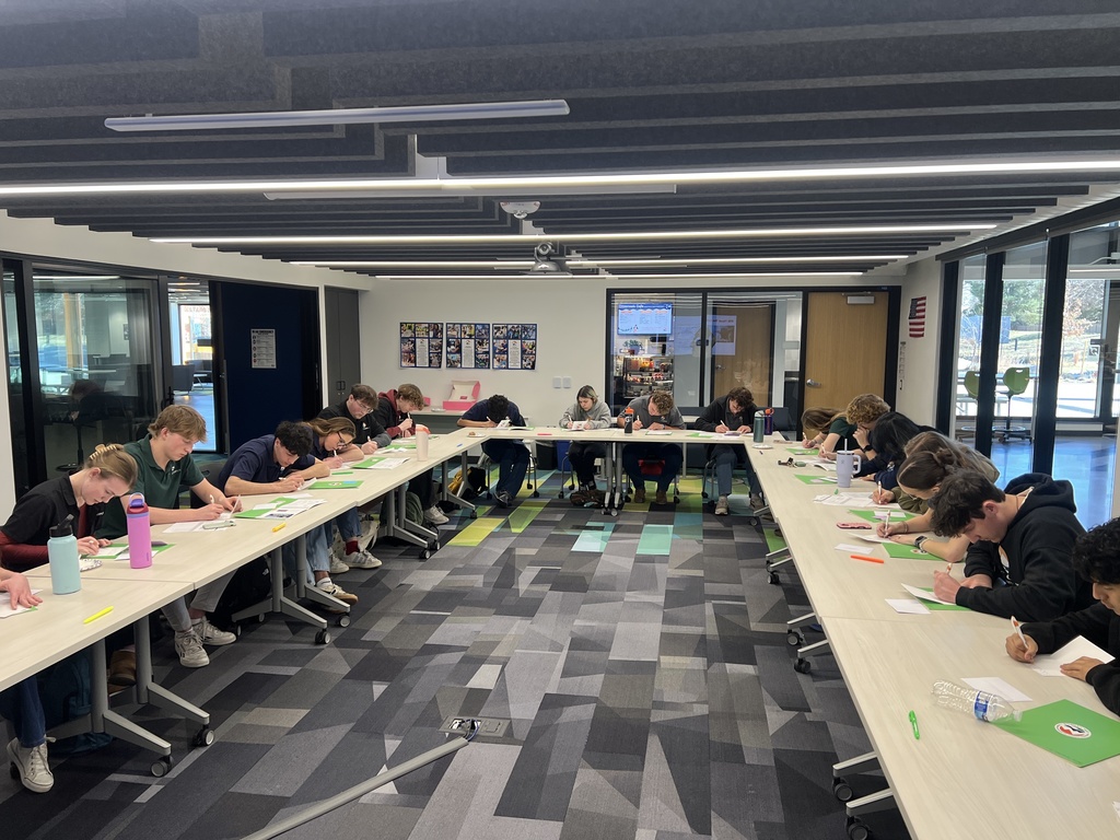 Students sit around a large U-shaped arrangement of white tables in a modern classroom, writing intently on paper. The diverse group of high school students includes individuals of various ethnicities and genders. Green folders, water bottles, and pens are spread across the tables. The room features colorful carpet tiles, large windows, glass walls, and an American flag on the far wall. A collage of photos and a digital screen are visible in the background.