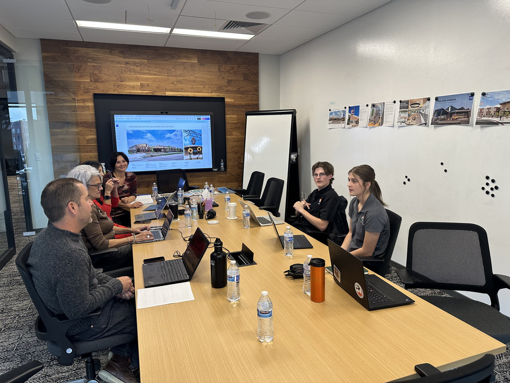 A diverse group of adults and students sit around a large conference table in a modern meeting room. They are engaged in discussion, using laptops and notebooks. A screen at the front displays a design rendering labeled “Spring Mountain Vision High School.” On the wall are printed images of architectural designs.