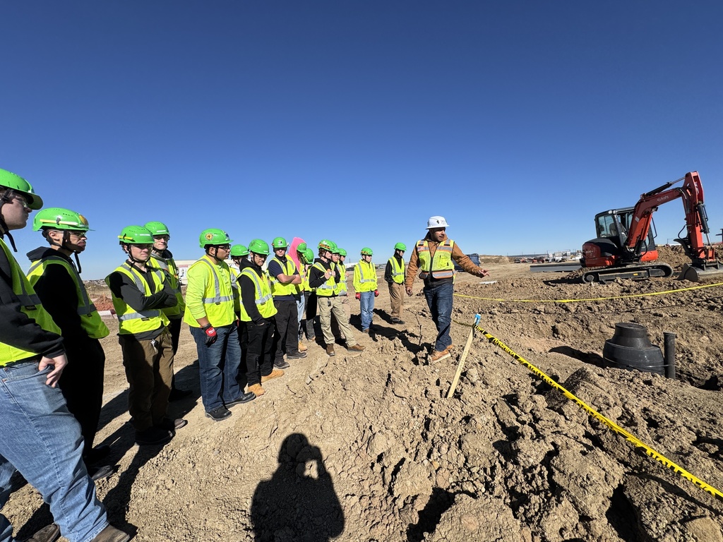 A group of students wearing green safety helmets and yellow vests stands at a construction site. An instructor in a white helmet and orange vest gestures toward the ground, marked off with caution tape. A red excavator is in the background under a clear blue sky.