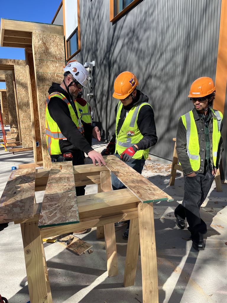 Two students in safety vests and hard hats receive instruction from an adult in construction gear outdoors. They are assembling a small wooden frame structure beside a modern building with wood and metal siding.