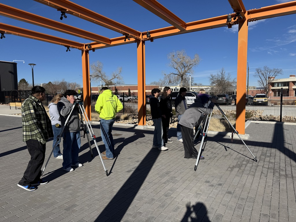 Several high school students participate in an outdoor surveying activity under a bright orange steel frame. Some are using tripods and survey tools, while others observe. The setting is a paved area near a school campus with buildings and cars in the background.