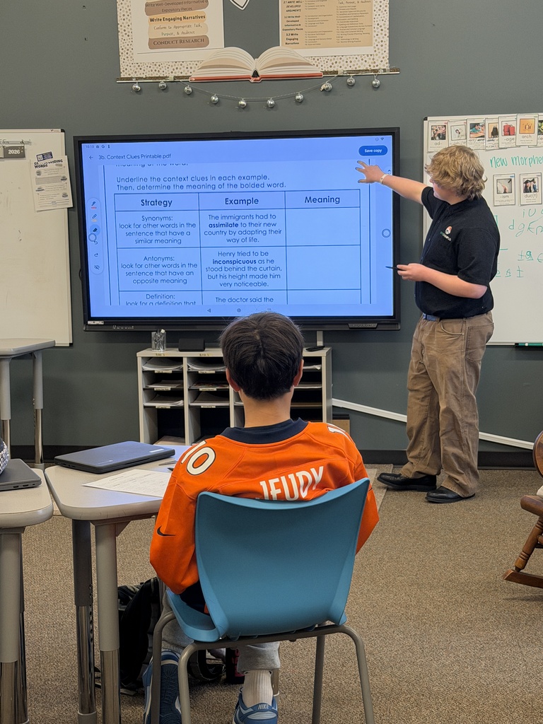A classroom scene with a student in a Broncos jersey seated and facing a large digital screen, while a teacher stands and points at the screen displaying a context clues worksheet. The student’s jersey reads “JEUDY 10.” The classroom includes modern furniture, laptops, and whiteboards with educational materials. The focus is on a learning activity involving reading strategies.