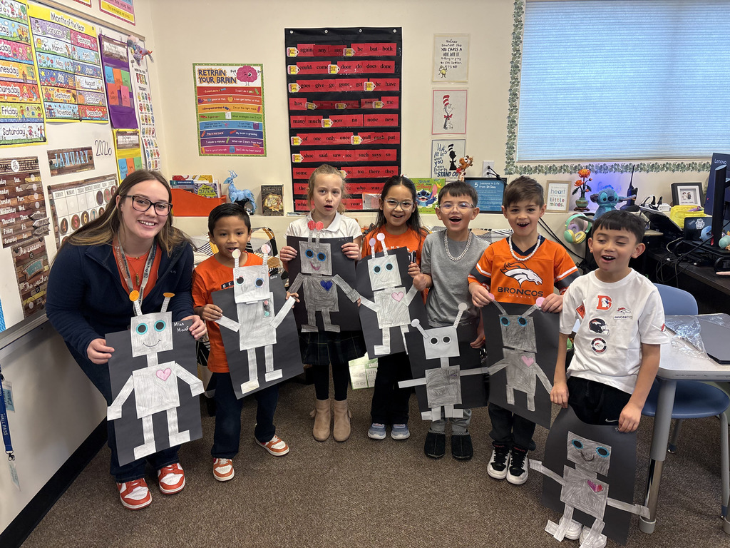 A group of eight elementary students and one teacher stand smiling in a classroom, holding paper robot crafts. The teacher and several students wear orange and black Broncos-themed clothing. The diverse group of children includes boys and girls of different ethnic backgrounds. The classroom is colorful with educational posters, a calendar showing January 2026, and a display of sight words on the wall behind them.