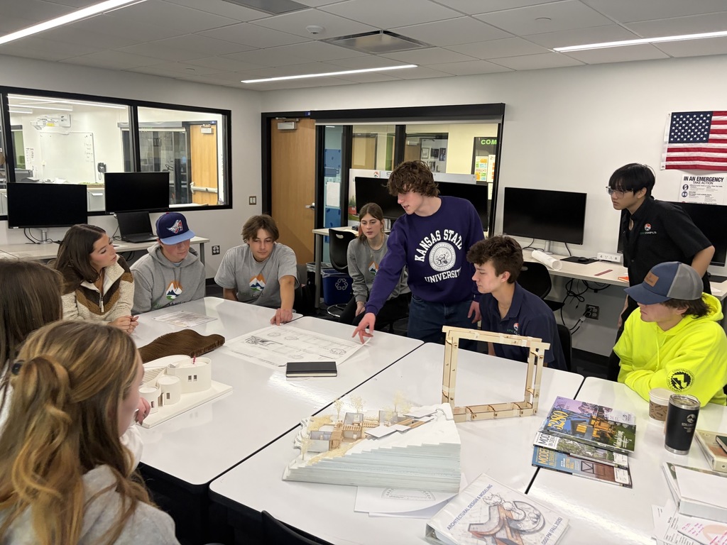 A group of high school students sit and stand around a large white table in a design or architecture classroom. They are attentively reviewing architectural plans and scale models placed on the table. One student in a Kansas State University sweatshirt gestures toward a drawing while others listen. The room features modern equipment, desktop monitors, and educational posters, including an American flag and safety signage on the wall.