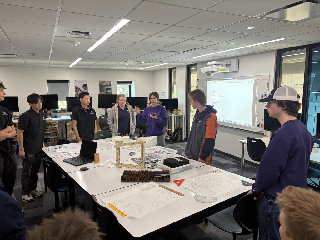 High school students stand around a table in a modern classroom, engaged in a group discussion. Architectural models, tools, and large design plans are spread across the table. The diverse group includes students of different genders and ethnic backgrounds. Some students wear matching black polo shirts with a logo, suggesting a team or class project. A digital whiteboard in the background displays a webpage, and multiple monitors line the back wall.