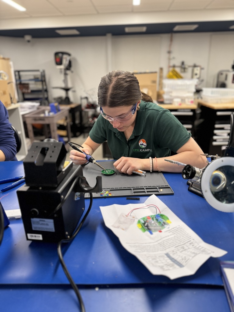 A student wearing safety goggles and a dark green EPIC Campus polo shirt uses a soldering iron to work on a green circuit board at a blue workstation. The student is surrounded by electronics tools and a printed guide with a diagram. The classroom is a well-equipped makerspace with visible workbenches and storage in the background.