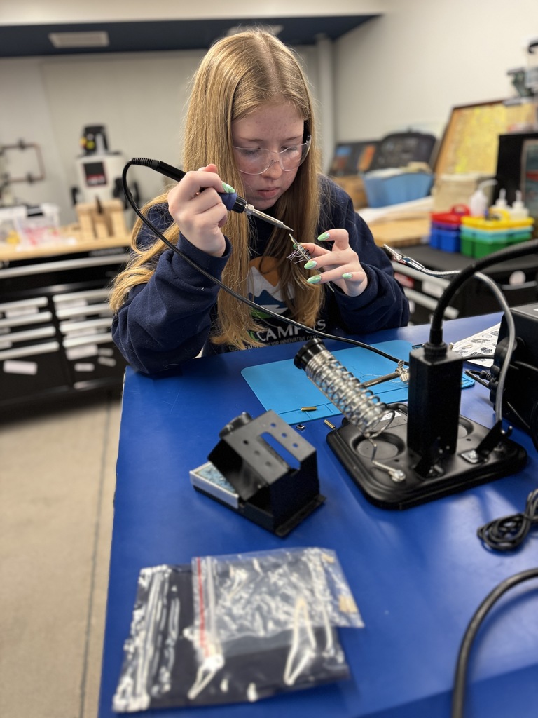 A high school student with long blonde hair wears safety goggles and a navy blue sweatshirt with the EPIC Campus logo while carefully using a soldering iron at a blue workbench. She focuses on connecting wires, with soldering tools and a bag of components on the table. The background includes organized drawers and classroom equipment, suggesting a hands-on STEM learning environment.