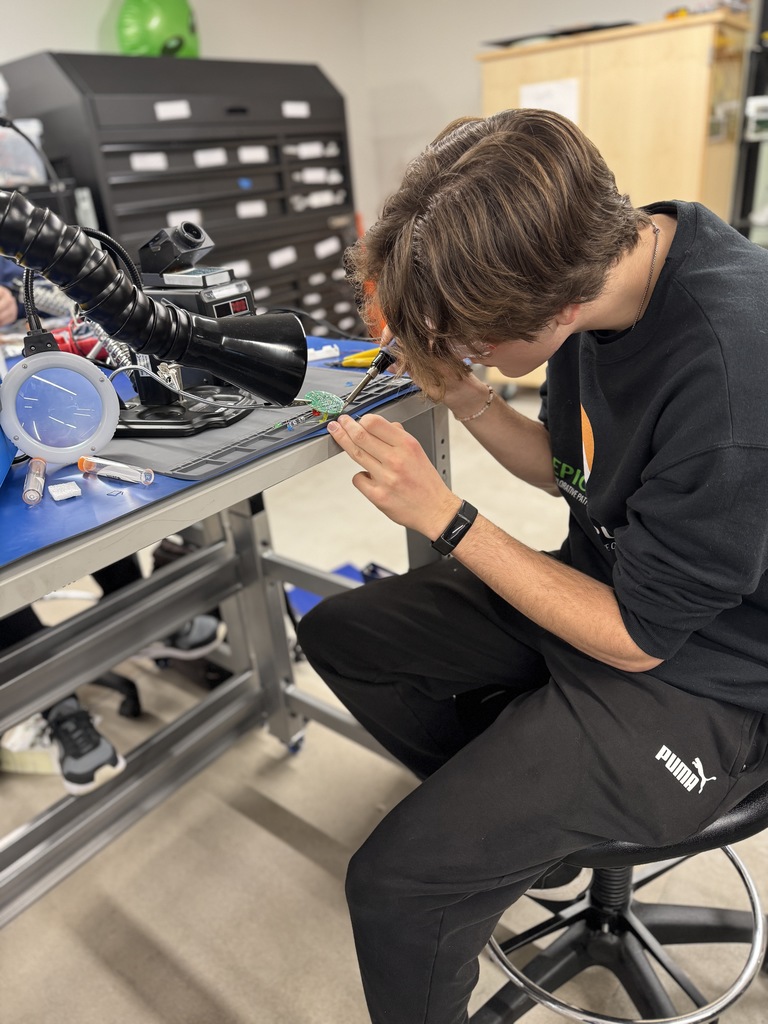 A student in a black EPIC Campus shirt and Puma pants leans over a table, using a soldering iron to work on a circuit board. He is seated on a stool in a well-organized tech classroom with tool drawers, a solder fume extractor, and various electronics equipment nearby.