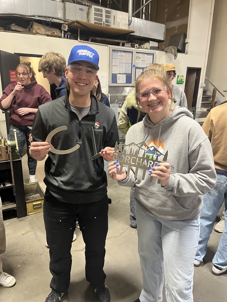 Two high school students wearing safety glasses smile and hold up custom-cut metal pieces inside a manufacturing workshop. The student on the left holds a large curved piece, and the student on the right displays a decorative cutout reading “ORCHARD” with a mountain design. Both wear EPIC Campus clothing, and the background shows classmates and industrial equipment.