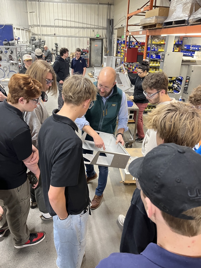 A group of high school students gathers around an instructor holding a large metal part inside a manufacturing workspace. The instructor points out features of the part while explaining, and the students observe closely. The workshop is filled with industrial shelving, tools, and metal components. Many students wear safety glasses, and one woman in the background works at a separate station.