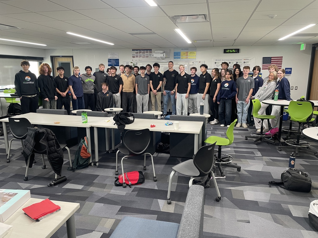 A diverse group of high school students stands in a line across a modern classroom, smiling at the camera. Most are wearing matching dark shirts with a "Team Bull" or LPS Capstone logo. The classroom features bright lighting, gray patterned carpet, whiteboards covered with charts, and U.S. and Colorado flags. Green and black chairs and scattered backpacks add to the casual academic setting.