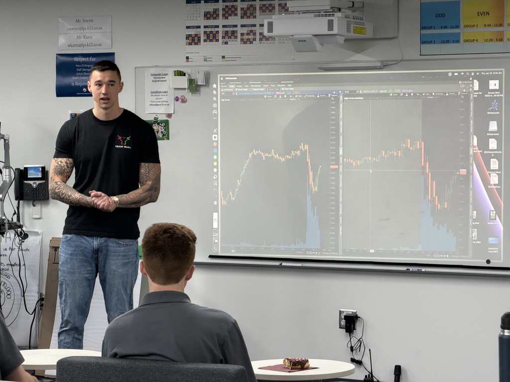A close-up view of the speaker presenting to students in a classroom. He is standing near a whiteboard with two large trading charts projected behind him. He wears a black "Team Bull" shirt with a red and green bull graphic and has full sleeve tattoos. A student with short hair is visible in the foreground, and a chocolate-frosted donut sits on the table.