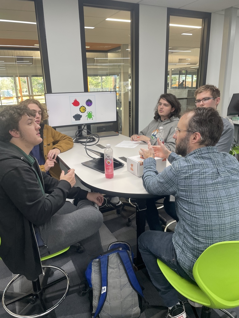 Five high school students and one adult sit around a circular table in a classroom, engaged in a discussion. A monitor displays colorful tiger paw logos in various styles. A laptop, drinks, and printed materials are on the table. The classroom has large interior windows, green chairs, and modern decor.