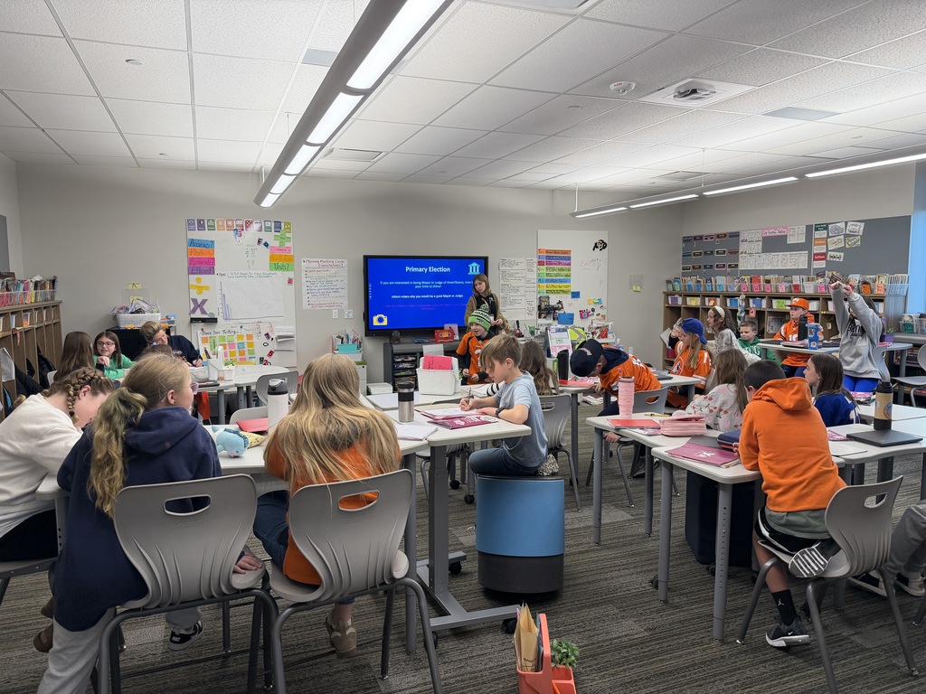 An elementary classroom full of students engaged in a lesson about primary elections, as shown on the screen at the front. The room is colorful and vibrant, with student work, posters, and vocabulary cards displayed on the walls. Most students are working at tables, while some raise their hands or look toward the front. Several students are wearing orange jerseys and hoodies.