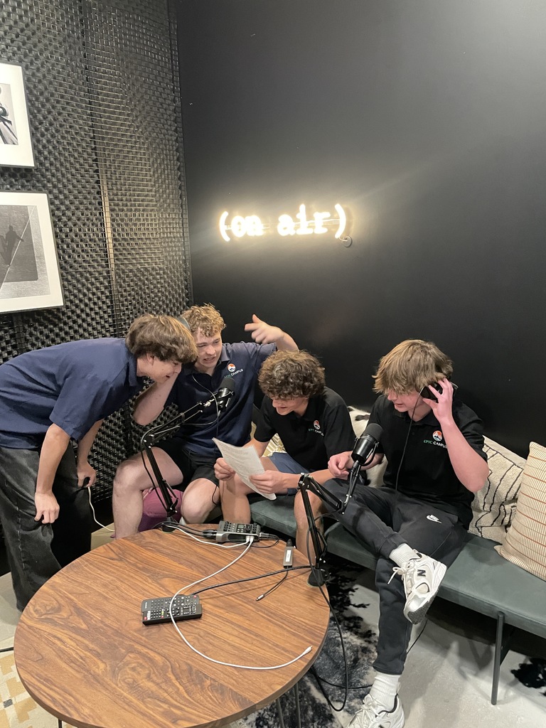 Four male students gather around microphones and a script in a podcast recording room. The group is engaged and smiling under a glowing “on air” sign. The space features acoustic wall panels and a cozy setup with a wooden table and tech equipment.