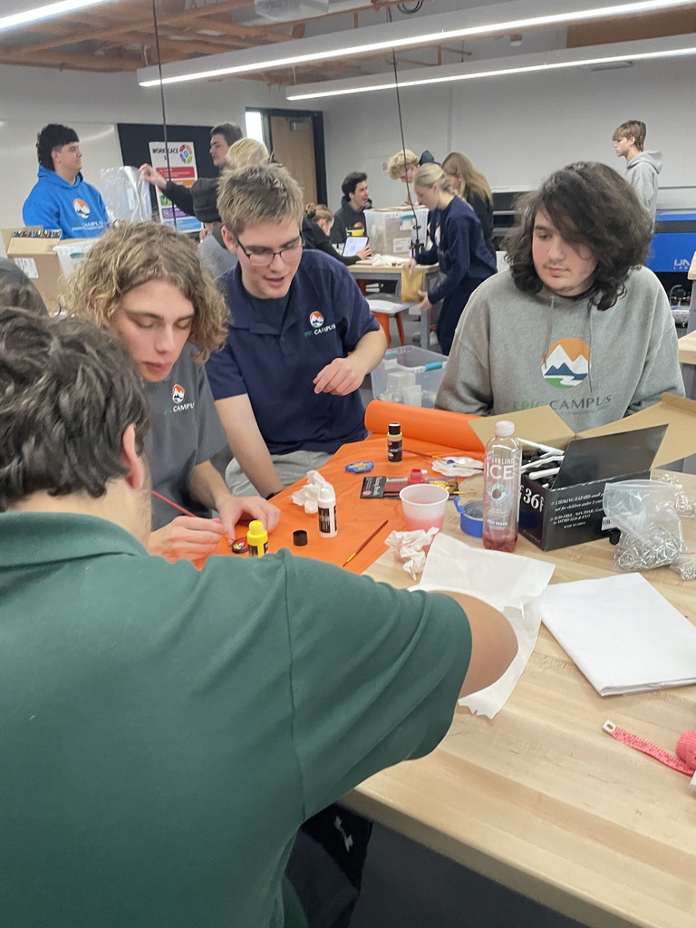A busy, hands-on classroom where a group of students work together at a table with tools, bottles, and craft supplies. Most students wear EPIC Campus sweatshirts or polos. The room has an industrial, maker-space style with students actively engaged in creative projects.
