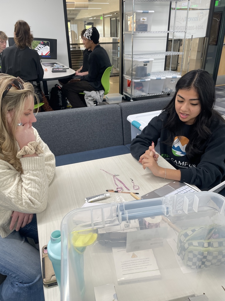 Two female students collaborate at a table with craft supplies, possibly working on jewelry or wire projects. One student wears an “EPIC Campus” sweatshirt. In the background, other students work at computers in a modern, open classroom.