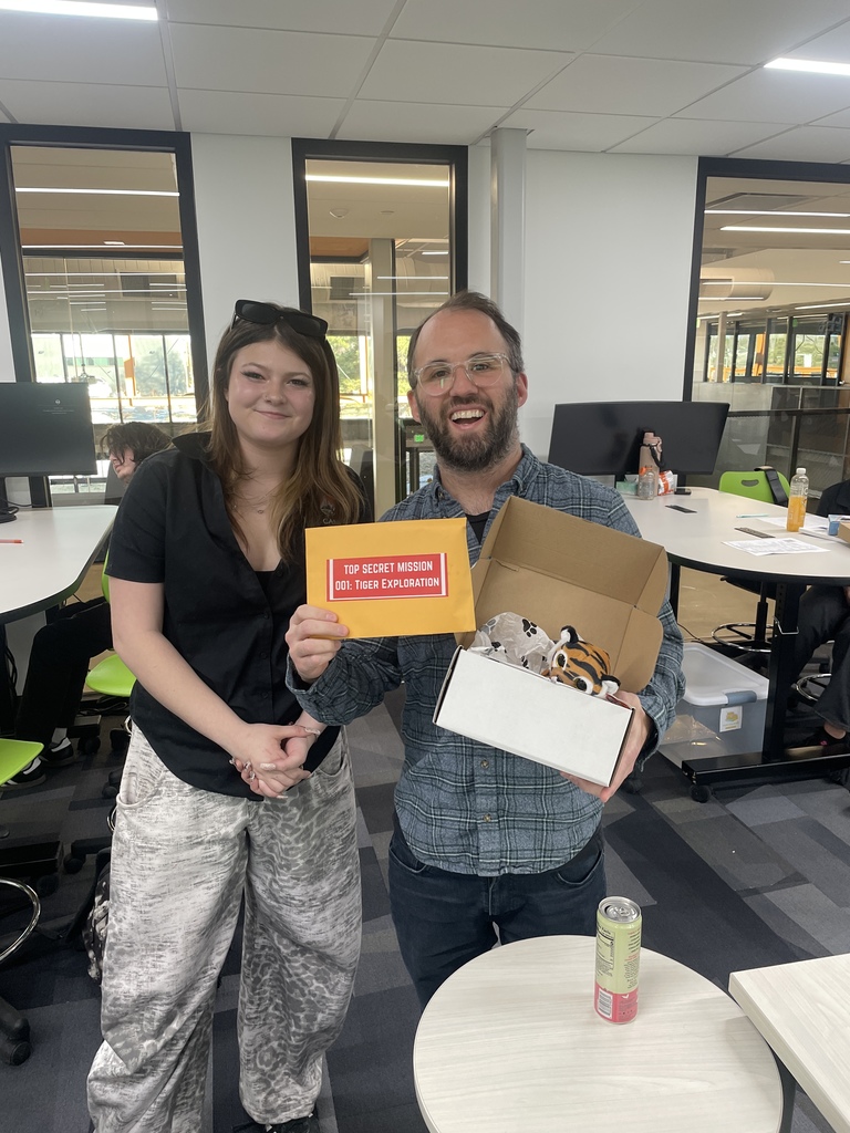 A man and woman stand smiling in a classroom. The man holds a box with a small plush tiger and a bright orange envelope labeled “Top Secret Mission 001: Tiger Exploration.” The environment is cheerful and modern with glass walls and green-accented chairs.