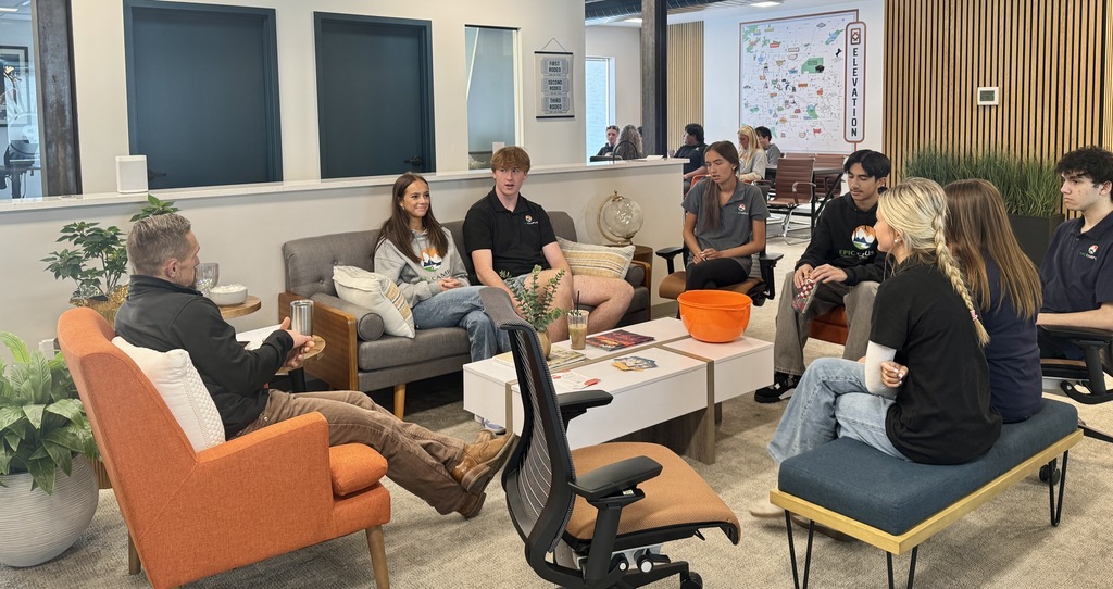 A casual discussion setting with students and an adult sitting on modern furniture in a collaborative workspace. The group includes a mix of male and female high school students, several wearing “EPIC Campus” shirts. The space features warm lighting, a mix of seating types, and a modern aesthetic.