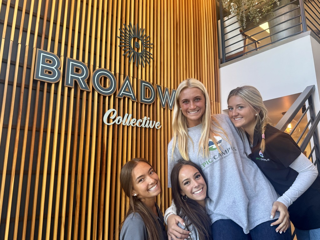 Four smiling female students pose in front of a wood-paneled wall with the words “Broadway Collective” in large letters. All are wearing “EPIC Campus” gear. A staircase and hanging plants are visible in the background, giving a modern and welcoming feel.