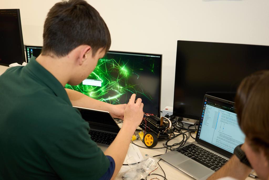 Two high school students collaborate at a desk with multiple monitors and laptops. One student in a green polo shirt works on wiring a small robotic car with yellow wheels, while the other reviews code on a laptop. The monitors display a neon green abstract background. The table is cluttered with wires and robotics equipment.