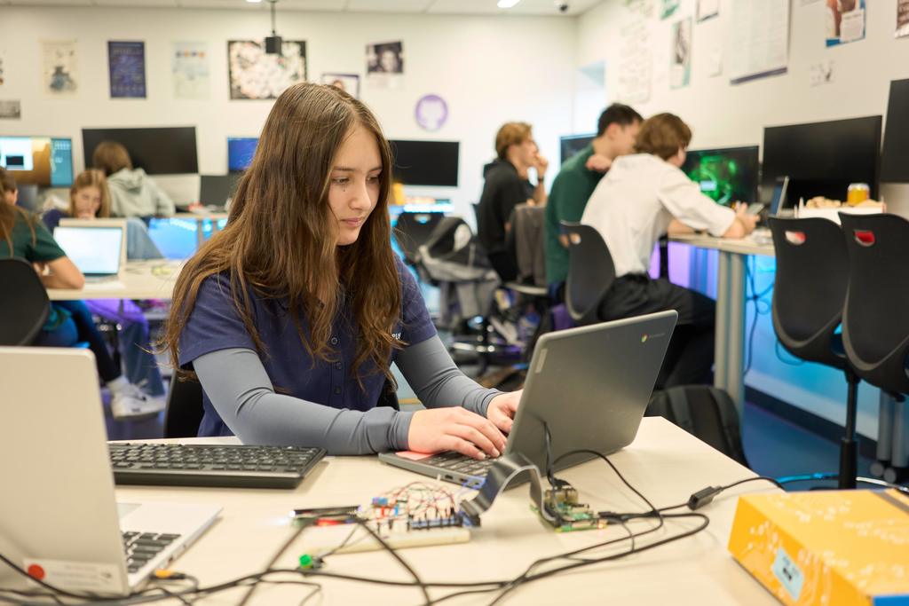A female high school student with long brown hair and a navy blue shirt types on a Chromebook at a classroom table. In front of her are wires and a small breadboard with electronic components. The background shows other students, mostly teens, engaged in computer work at standing desks with glowing LED lights. Classroom walls are decorated with posters.