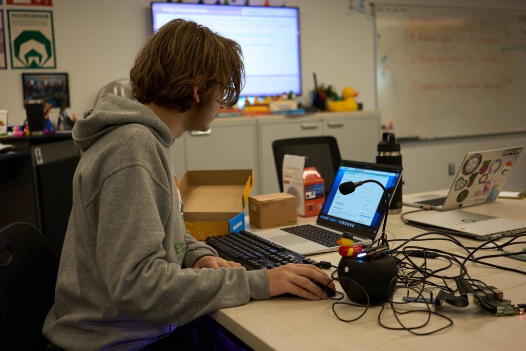 A high school student with shoulder-length brown hair, wearing a gray hoodie, works at a table with a laptop, external keyboard, and various tech components including a microphone, wires, circuit boards, and a rubber duck. Multiple cardboard boxes are on the table. Behind him is a whiteboard, classroom posters, and a large display screen showing digital content. The room has a STEM lab feel.