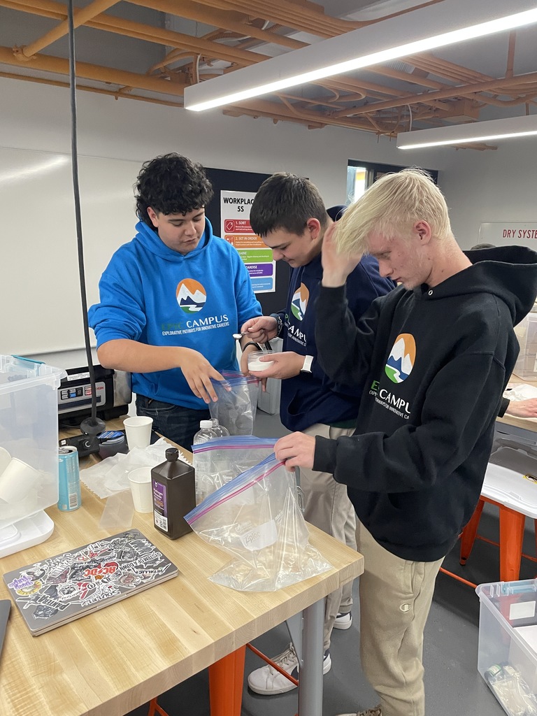 Three high school boys stand around a table working with plastic bags and white powder, likely for a hands-on science or engineering activity. One wears a blue EPIC Campus hoodie and the others wear black. A laptop covered in stickers and other materials are visible on the table.