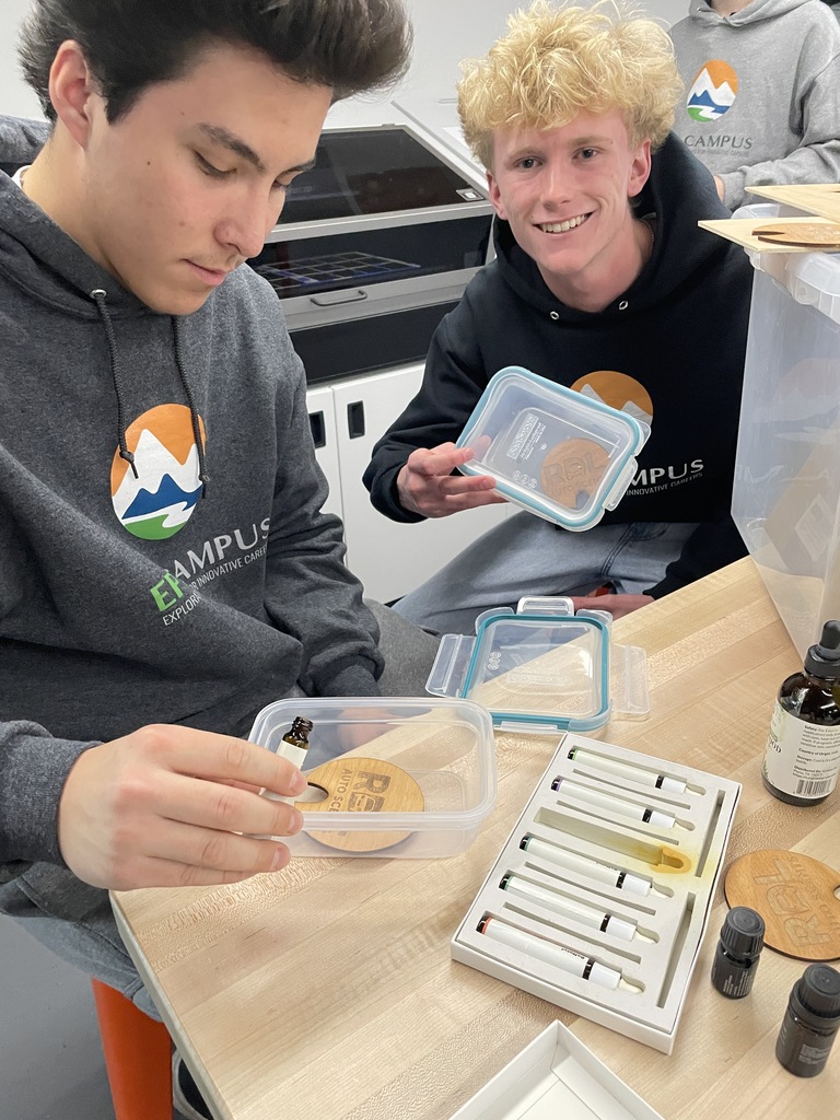 Two male students work with a set of essential oil droppers and wooden coasters labeled "REZ." One student is smiling at the camera and holding up a plastic container with a wooden piece inside. The other focuses on applying oil. Both wear EPIC Campus hoodies in a bright classroom lab.