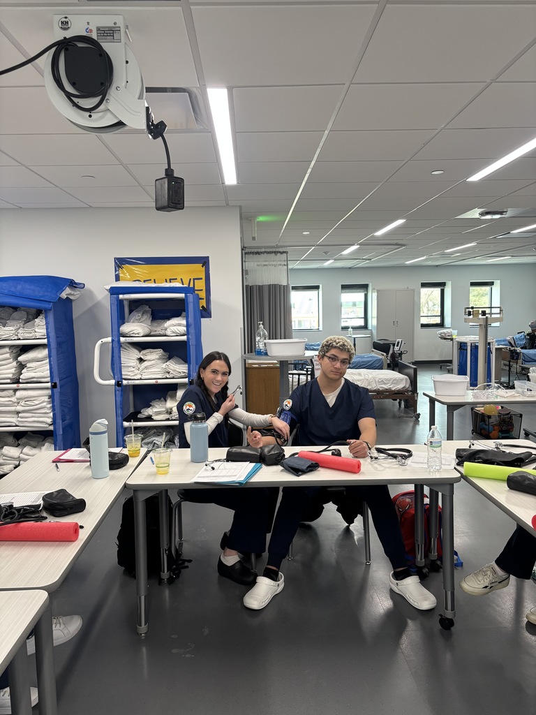 Two students in navy blue medical scrubs sit at a classroom table practicing blood pressure readings with a cuff and stethoscope. They are smiling and interacting, surrounded by water bottles, clipboards, and medical supplies. In the background are neatly stacked shelves with folded linens and a sign that reads “BELIEVE.”