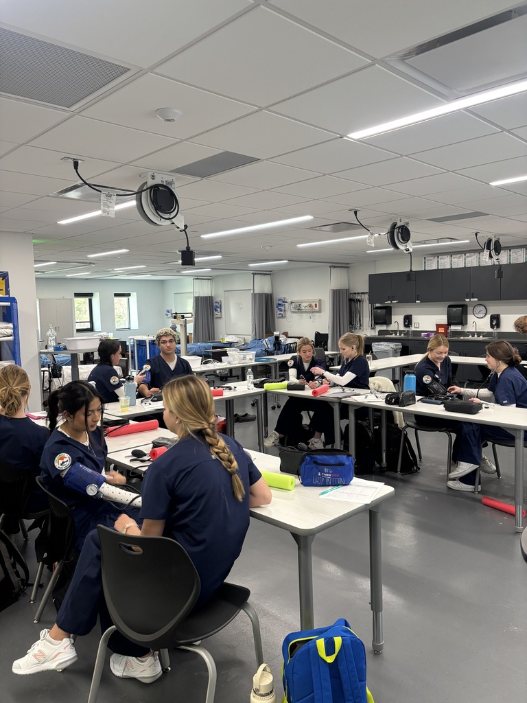 A large classroom with students in matching navy scrubs practicing clinical skills in pairs. Most students are using blood pressure cuffs and stethoscopes at white tables. The room is equipped with medical beds, cabinetry, and wall-mounted supplies. Overhead, retractable power reels and bright lighting are visible.