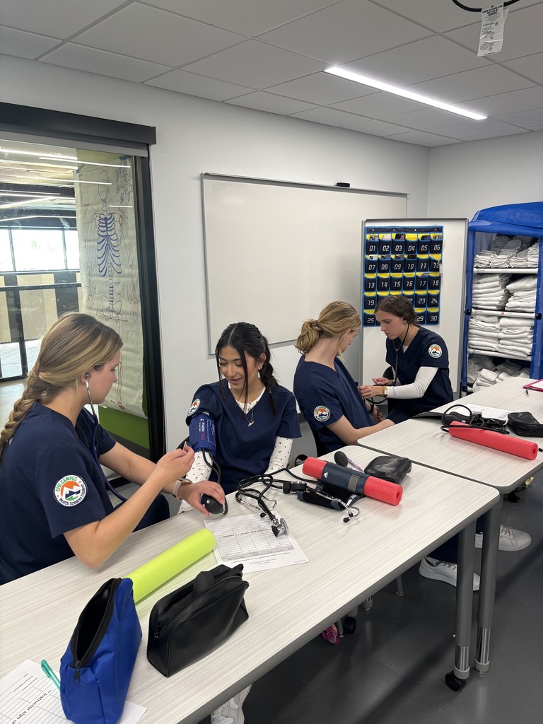 Four female students in navy blue scrubs pair up to practice taking each other's blood pressure. Each pair sits at a table with medical equipment, papers, and foam rollers. Behind them are shelves with linens and a whiteboard. One student uses a manual cuff and stethoscope while the other looks on attentively.