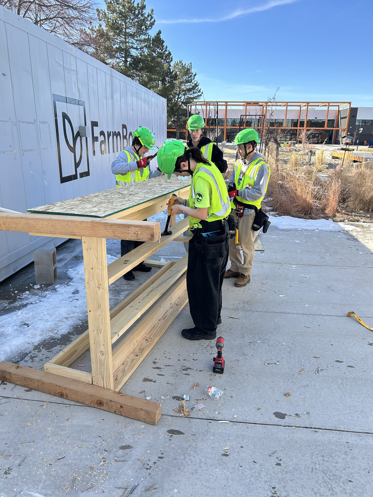 Five students in safety vests and green helmets work together on assembling a wooden bench or framework near a white shipping container labeled “FarmBox.” They are using hammers and drills, surrounded by sawdust and building materials. In the background is a modern building and landscaping with dried grasses.