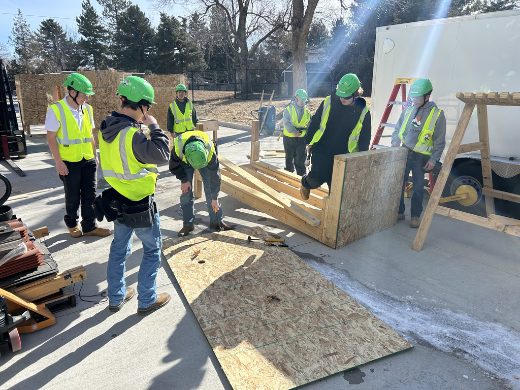 High school students wearing neon yellow safety vests and green hard hats work outdoors on a construction project. They are gathered around a partially built wooden frame structure, with one student hammering while others observe or assist. The scene is sunlit with visible trees, a trailer, and construction tools nearby.