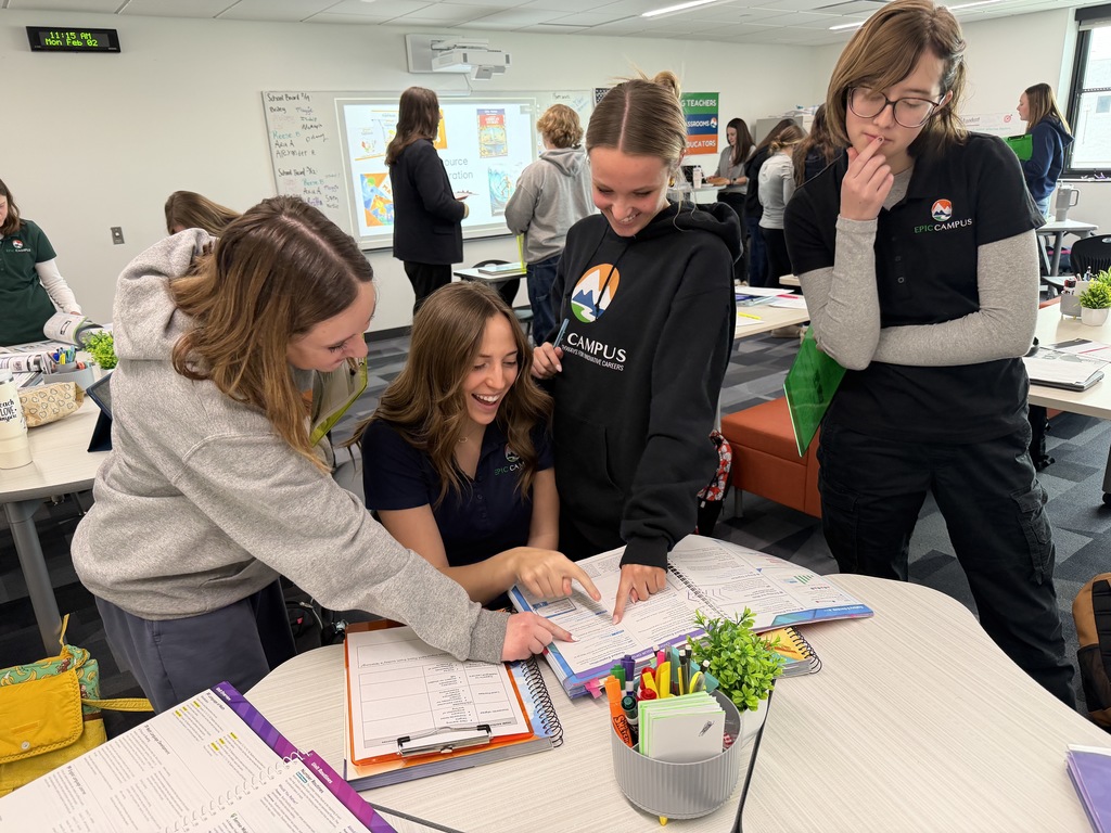 Four high school students stand around a table looking at a workbook together, smiling and pointing at something on the page. They wear EPIC Campus shirts and hoodies, and the table is filled with supplies like pens, binders, and a small plant. The mood is collaborative and cheerful.