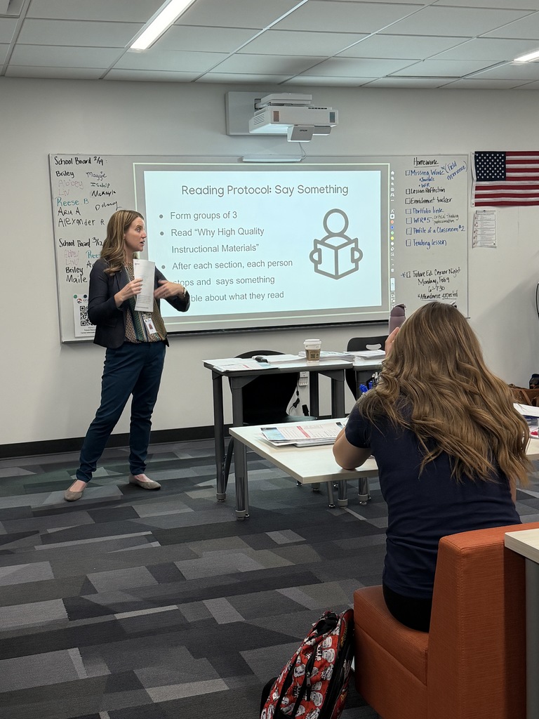 A female instructor stands in front of a projected slide guiding a reading protocol while a student listens attentively. The whiteboard beside the projector has handwritten notes and student names. The classroom has clean, modern decor with gray checkered carpet and white tables.