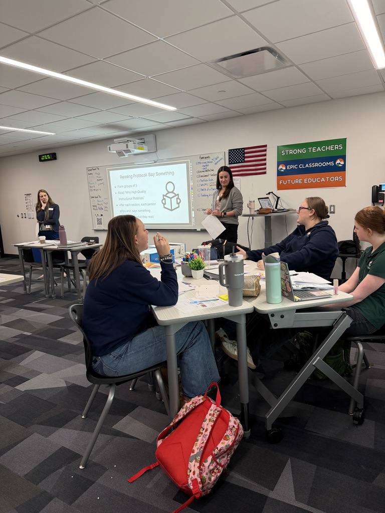 A high school classroom with students seated at group tables while a female instructor presents at the front near a projected slide titled "Reading Protocol: Say Something." Students are engaged in discussion, with notebooks, laptops, and water bottles on the tables. The room has an American flag, colorful signage on the wall promoting future educators, and a visible clock.