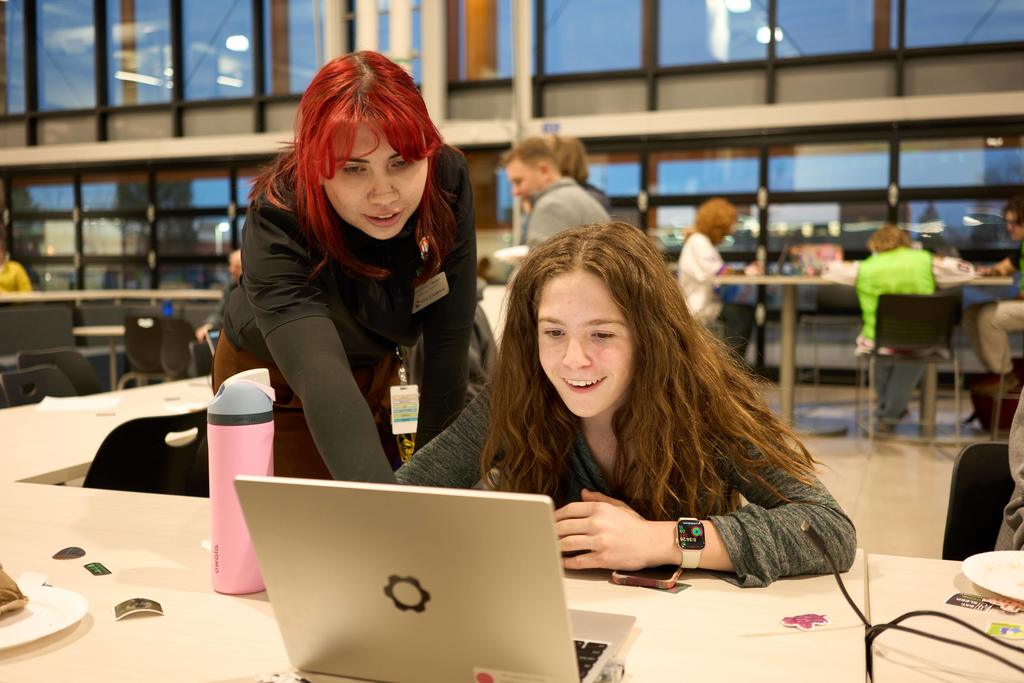 A student with long brown hair smiles at a laptop while an adult with red hair and a staff badge leans over to assist. They are seated at a table in a bright, modern common area with large windows and other students working in the background.