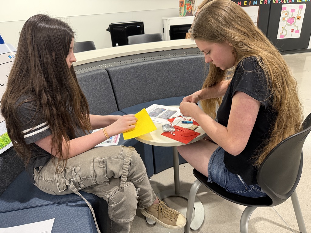 Two girls sit at a small round table, working on a craft project involving felt, thread, and paper. One student sews with red fabric while the other folds a piece of yellow material. They are seated in a bright, open learning space with educational posters on the wall.