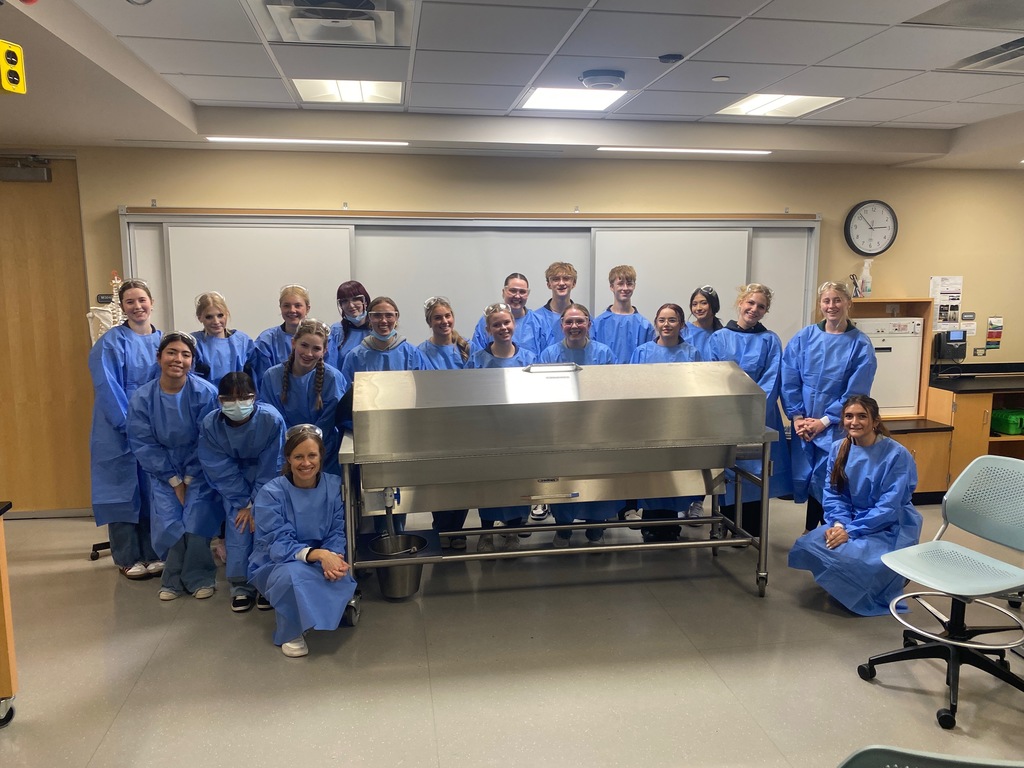A group of about 20 students and one adult stand together in a classroom around a large metal medical table. All are wearing blue surgical gowns and smiling at the camera. The room has lab equipment, a clock on the wall, and bright overhead lighting.