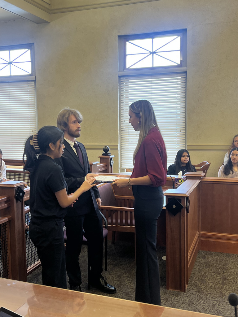 Three students stand and talk in a mock courtroom setting. Two female students and one male student are dressed in business attire, holding a book titled “LAW ENFORCEMENT.” Other students are seated in jury box-style benches in the background, watching the interaction.