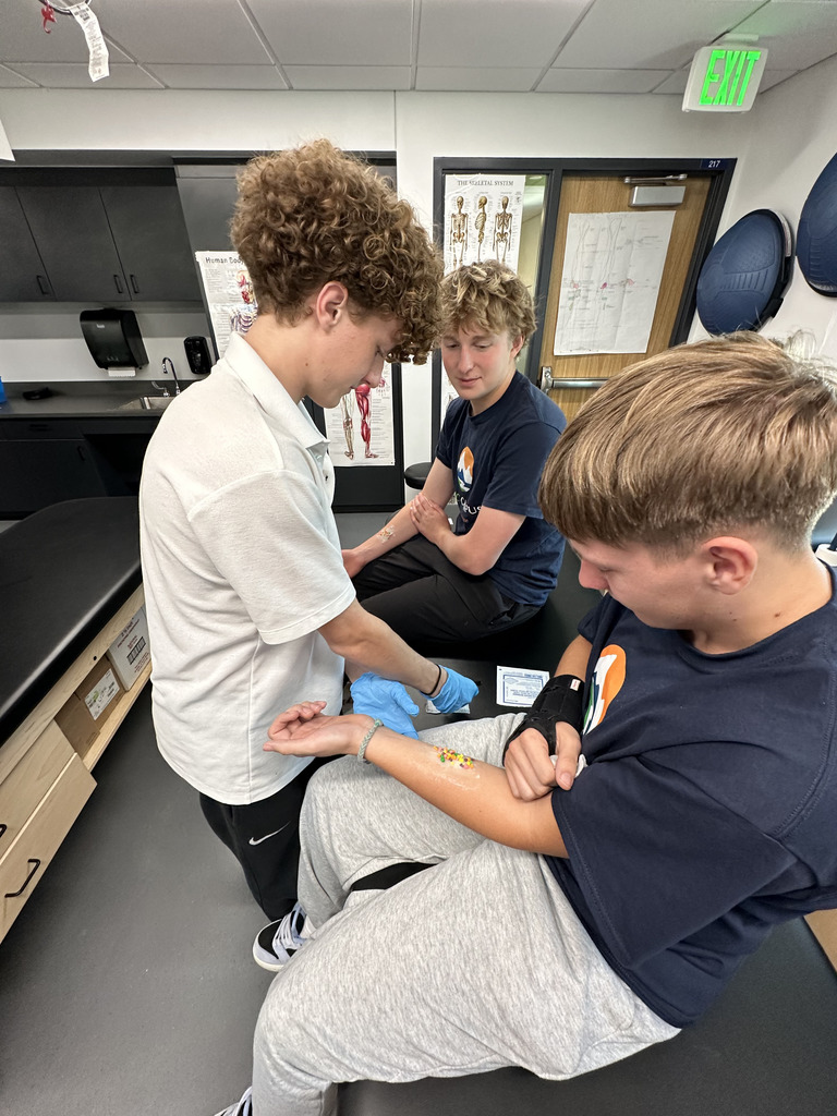 Three high school students in a medical classroom setting practice wound care. One student wears gloves and applies treatment to a mock injury on another student’s forearm, which is covered in simulated blood and medical dressing. A third student observes. Anatomical posters and diagrams are visible in the background.