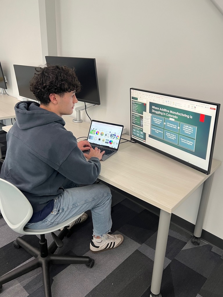 Student seated at a desk using a laptop connected to a large monitor. The screen displays a presentation titled “Where Additive Manufacturing Is Struggling in Colorado.” Additional computer monitors and a clean workspace are visible.